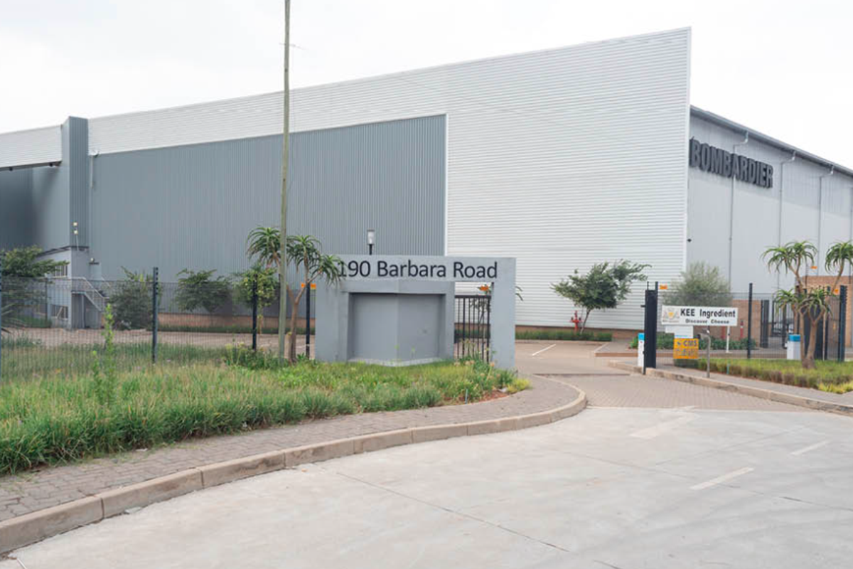Front exterior view of a large industrial warehouse facility featuring metal cladding, secure perimeter fencing, and a paved entrance driveway.