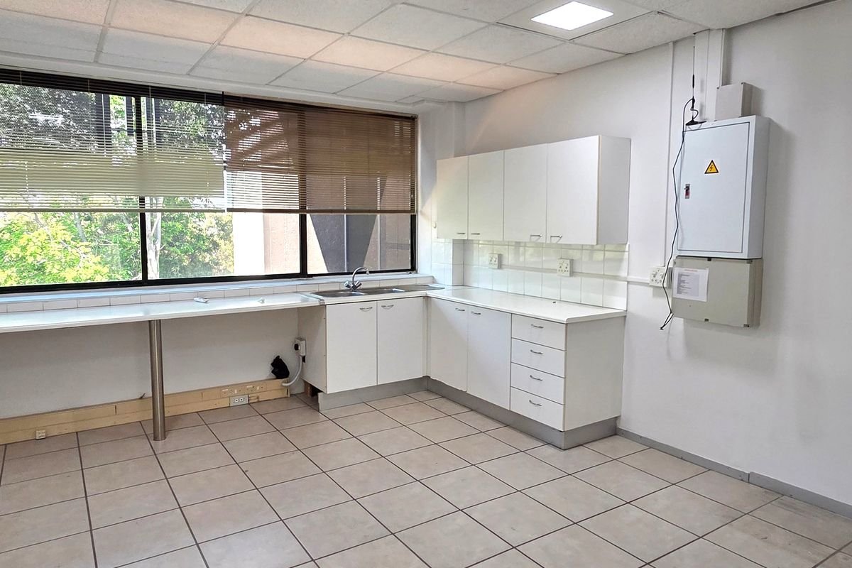 A functional office kitchenette featuring white built-in cabinetry, a stainless steel sink, tiled flooring, and a suspended ceiling with integrated lighting.