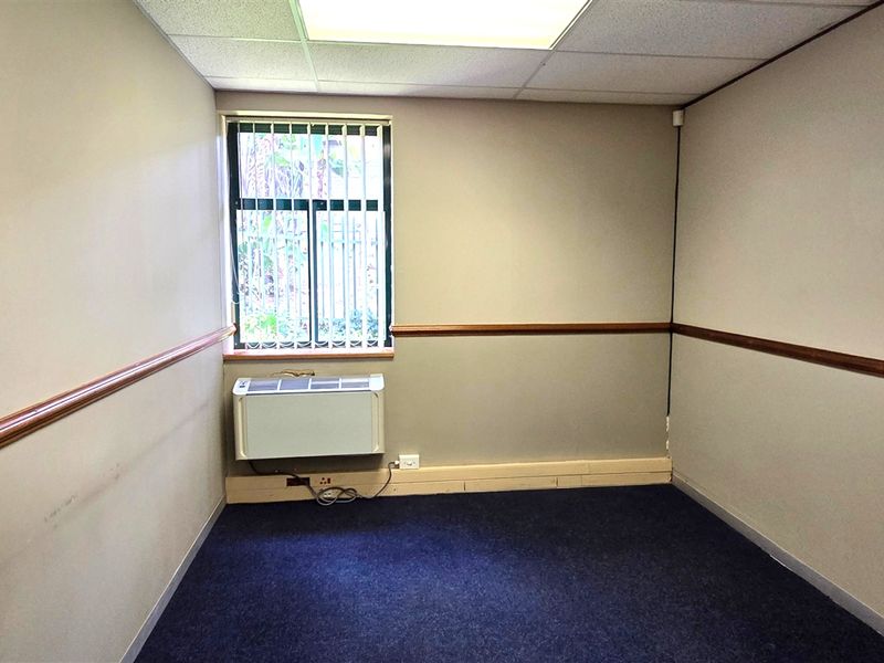 A vacant office space featuring carpeted flooring, a wall-mounted air conditioning unit, and a window with security bars.