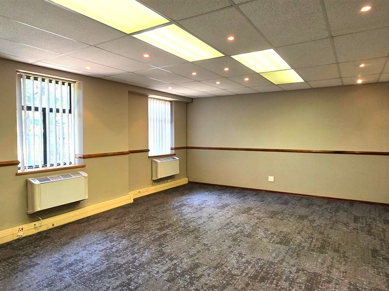 A spacious, empty office featuring carpet tiles, suspended ceilings with integrated lighting, wall-mounted HVAC units, and large windows with vertical blinds.