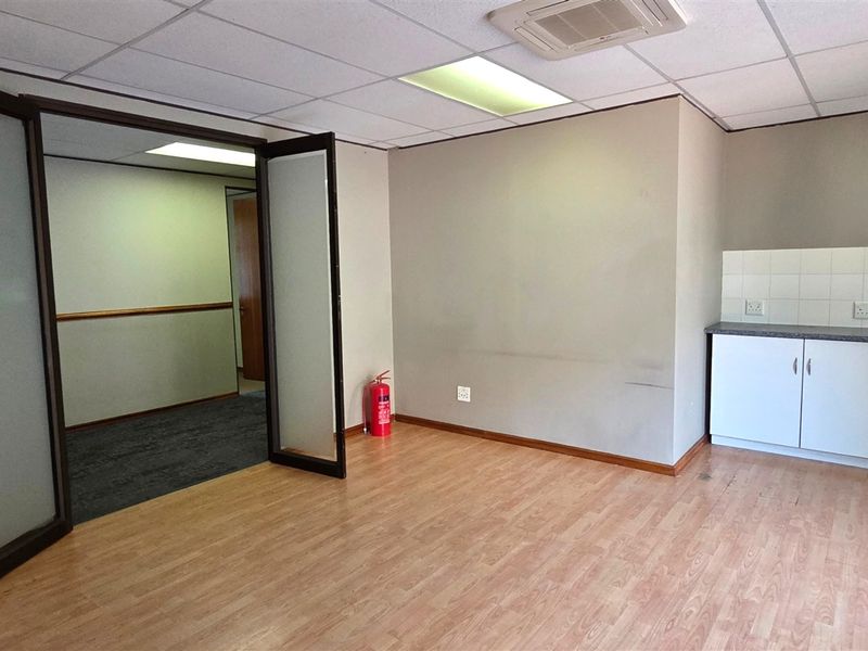 An open-plan office space featuring laminate flooring, a suspended ceiling with recessed lighting, a glass partition door, and a small kitchenette area.