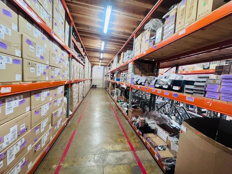 Industrial warehouse storage area featuring extensive steel racking systems, concrete flooring, and overhead fluorescent lighting.
