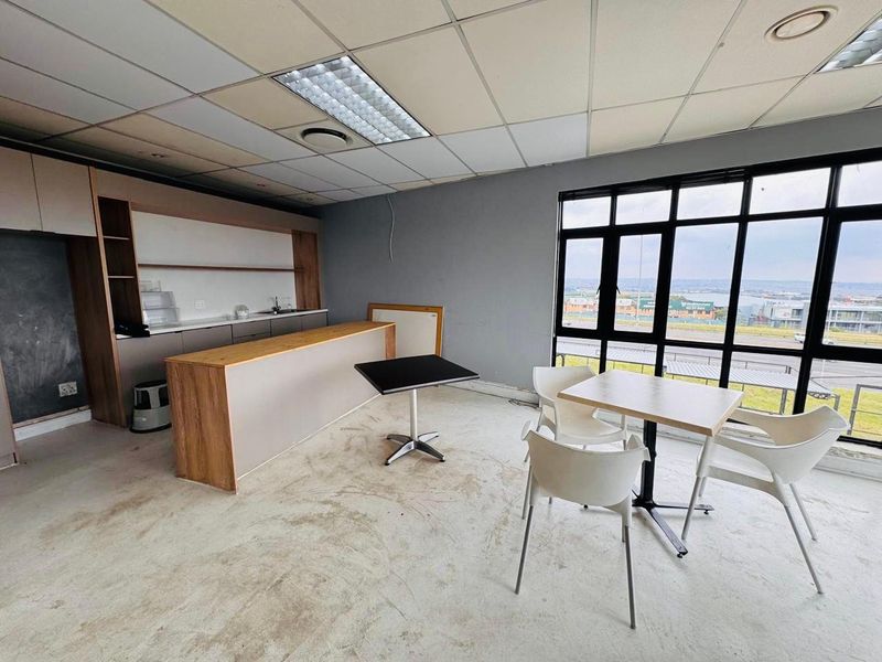 Open plan industrial office space featuring a kitchenette, drop ceiling, and large windows with exterior views. Requires flooring and ceiling maintenance.