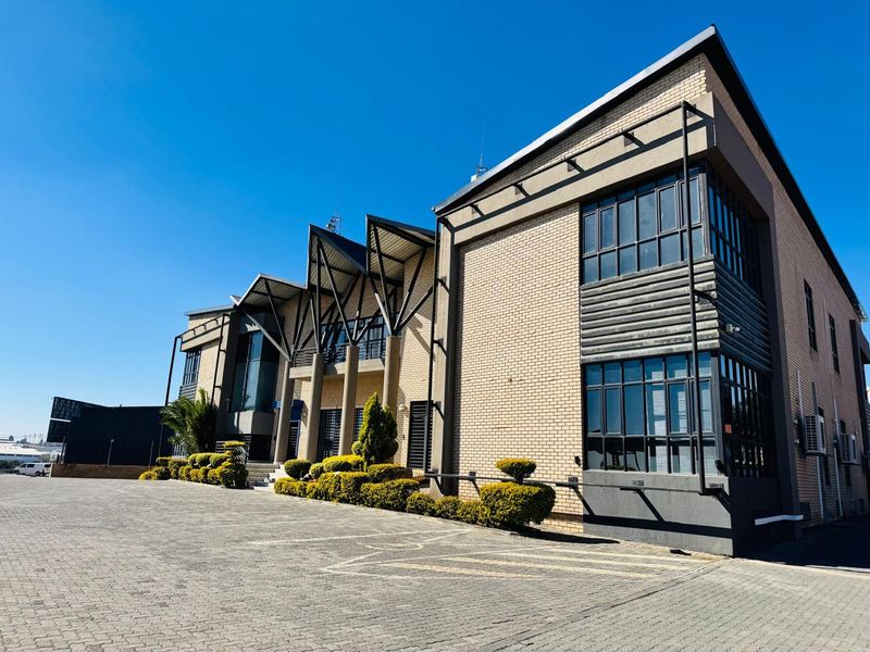 Modern industrial office building featuring a brick facade, prominent steel roof architectural elements, and extensive glass windows with a paved front parking area.