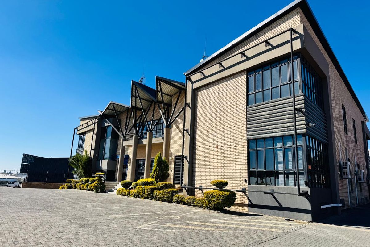 Modern industrial office building featuring a brick facade, prominent steel roof architectural elements, and extensive glass windows with a paved front parking area.