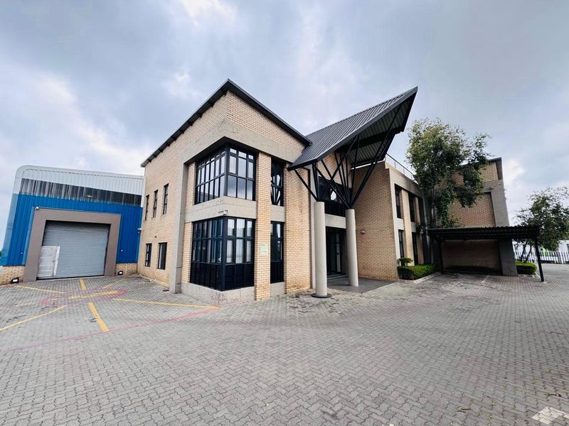 Modern industrial office building featuring a brick facade, large windows, a prominent covered entrance, and a paved driveway with a roller shutter door.