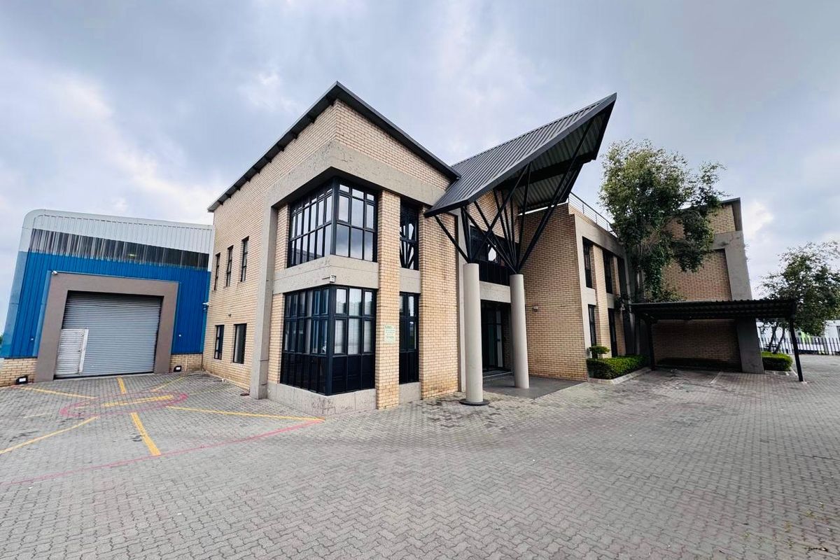 Modern industrial office building featuring a brick facade, large windows, a prominent covered entrance, and a paved driveway with a roller shutter door.