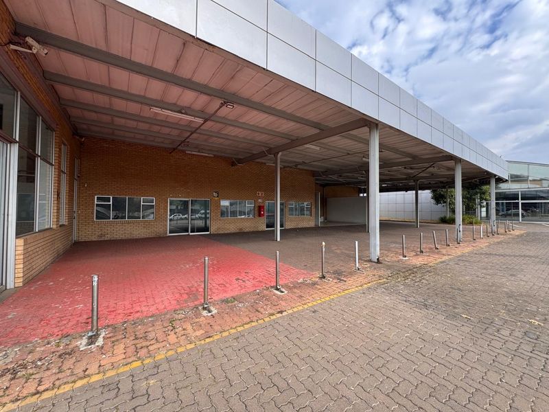 A commercial retail exterior featuring a large covered loading bay, brick facade, and paved driveway with protective steel bollards.