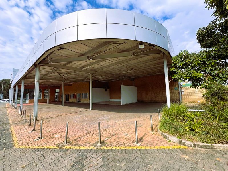 A commercial retail property exterior featuring a large covered canopy area, brick facade, and paved frontage with protective steel bollards.