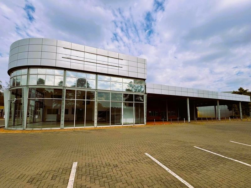 Modern commercial retail building featuring a prominent curved glass facade, metal panel cladding, and an expansive paved parking area with a covered canopy.
