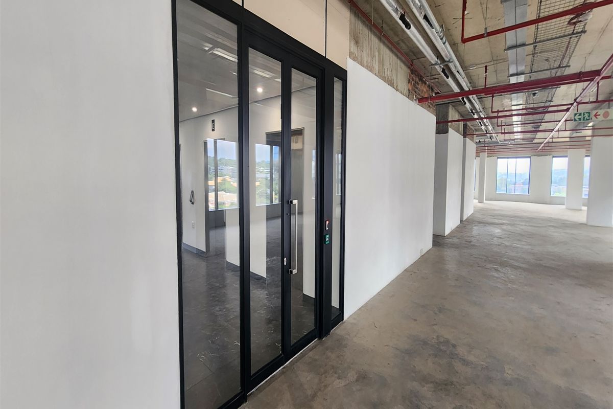 An open-plan office space featuring exposed industrial ceiling services, concrete flooring, and a modern glass partition entrance.