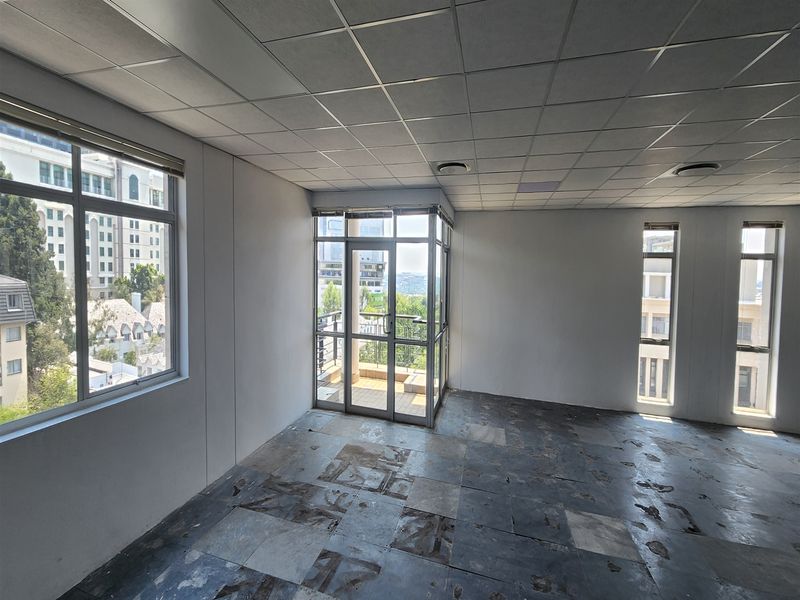 An empty office space featuring a suspended ceiling, large windows with natural light, and access to a small balcony. Requires flooring renovation.