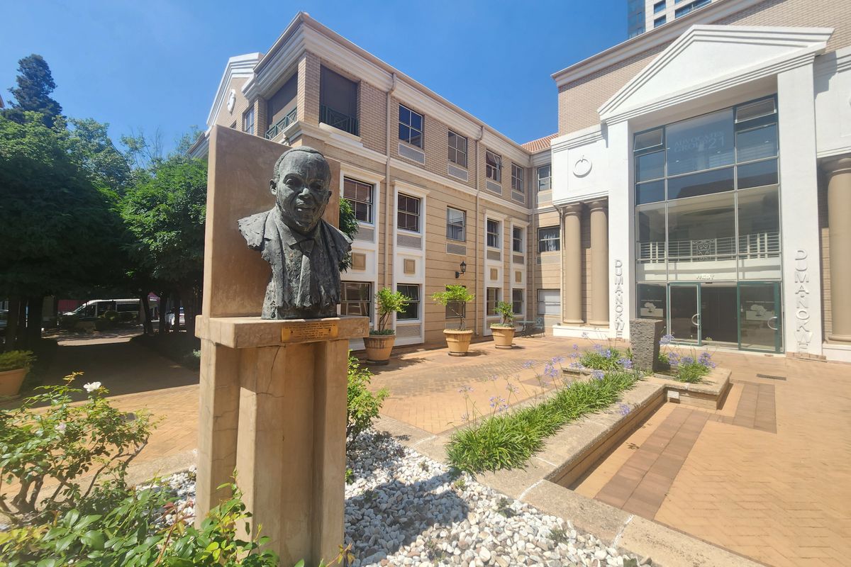 A well-maintained office building courtyard featuring brick paving, landscaped garden beds, and a prominent stone sculpture at the entrance.