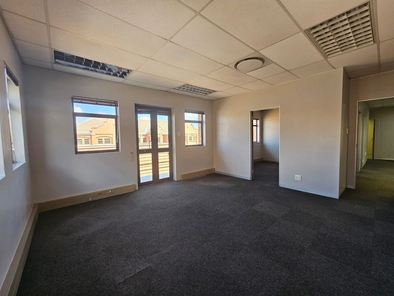 An empty, carpeted office space featuring a suspended ceiling, recessed lighting, and double glass doors leading to an exterior area.