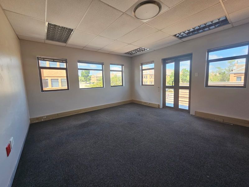 An empty, well-lit office space featuring carpeted flooring, suspended ceilings with recessed lighting, and multiple windows providing natural light.