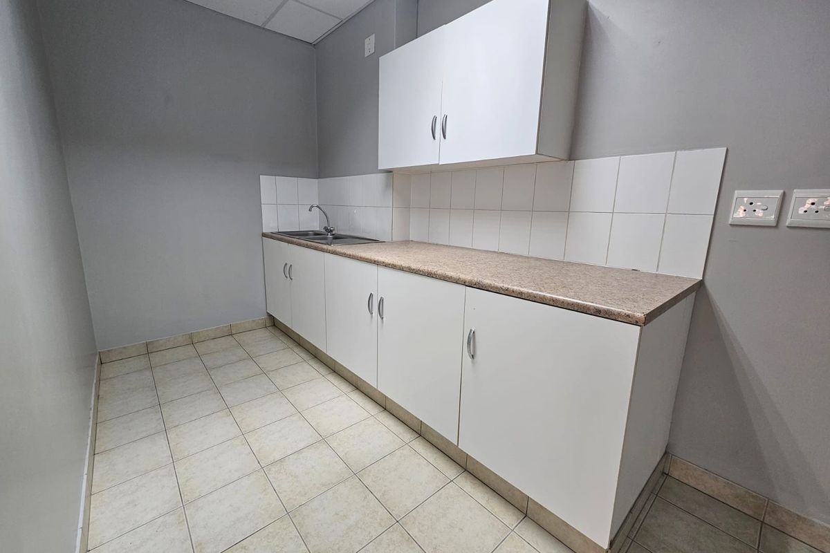 A functional kitchenette featuring white built-in cabinetry, a laminate countertop, stainless steel sink, and tiled splashback in a neutral office space.