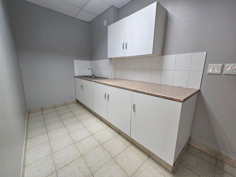 A functional kitchenette featuring white built-in cabinetry, a laminate countertop, stainless steel sink, and tiled splashback in a neutral office space.