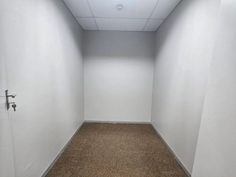 A compact, empty office space featuring neutral walls, carpeted flooring, and a suspended ceiling with an integrated smoke detector.