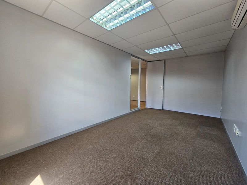 A vacant, functional office space featuring neutral carpeting, a suspended ceiling with recessed lighting, and a split-system air conditioner.