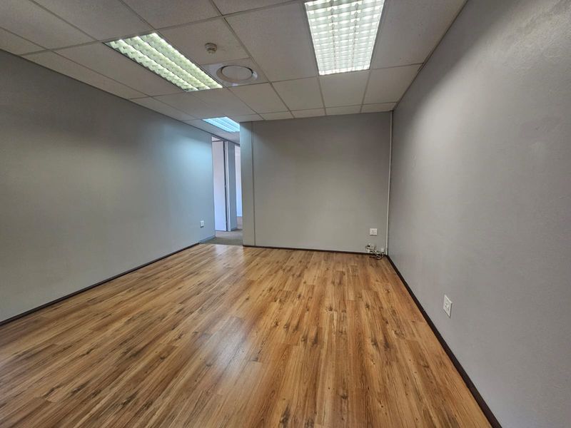 A clean, empty office space featuring wood-look laminate flooring, a suspended ceiling with recessed lighting, and neutral grey walls.