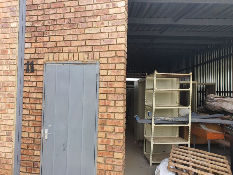Industrial unit entrance featuring exposed brickwork, a metal door, and an open-plan storage or utility area with corrugated metal roofing.