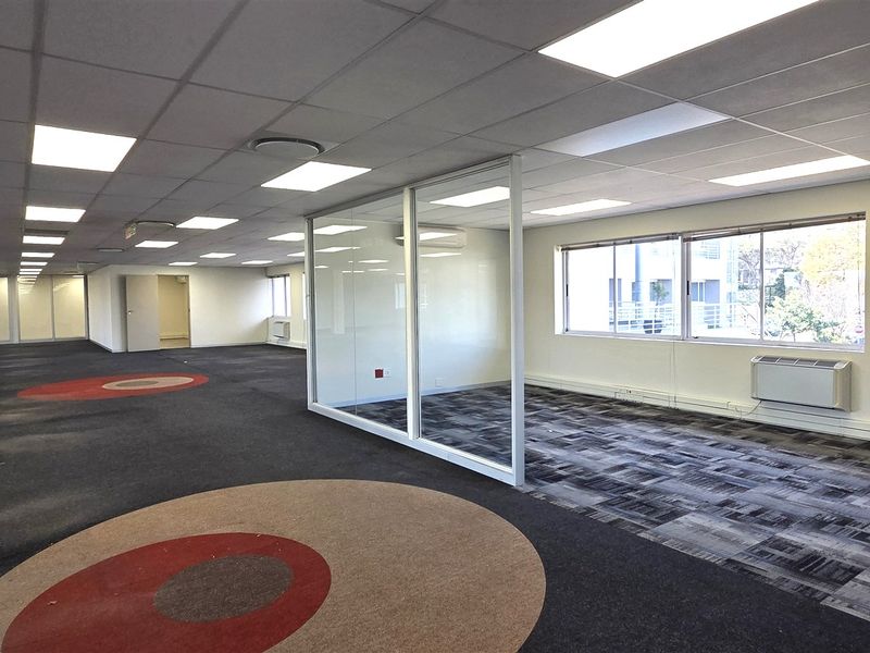 Spacious open-plan office area featuring a glass-partitioned private office, suspended ceilings with recessed lighting, and carpeted flooring throughout.
