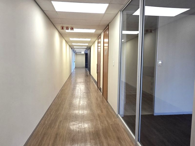 A clean, well-lit office hallway featuring laminate flooring, suspended ceilings with recessed lighting, and glass partition walls leading to individual office spaces.