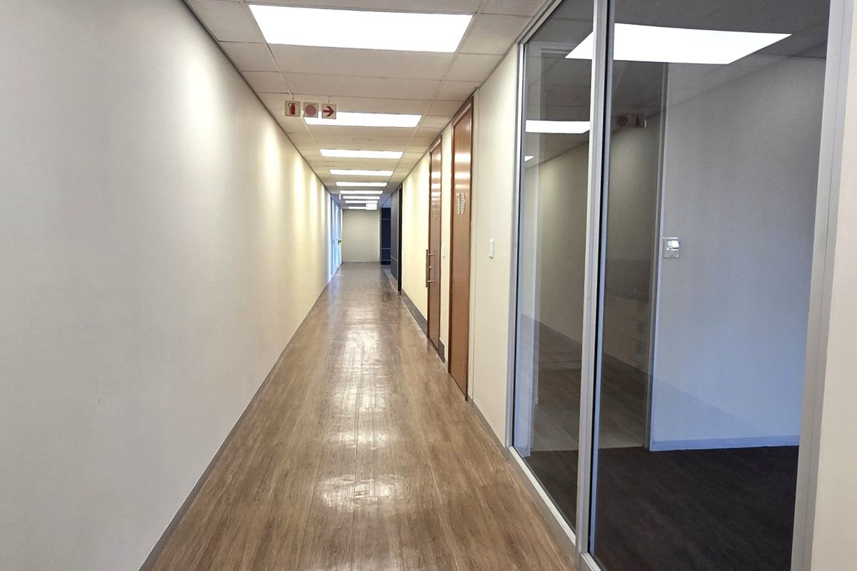 A clean, well-lit office hallway featuring laminate flooring, suspended ceilings with recessed lighting, and glass partition walls leading to individual office spaces.