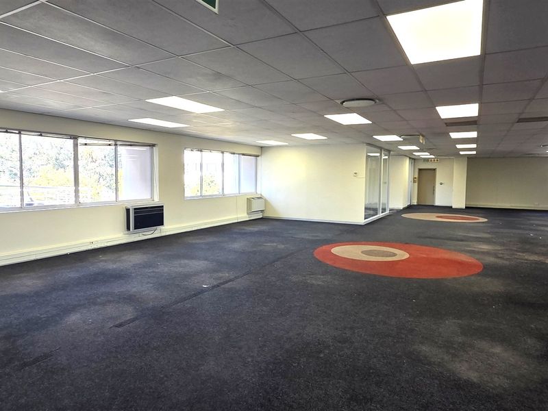 Spacious open-plan office area featuring suspended ceilings, recessed lighting, wall-mounted air conditioning units, and large windows providing ample natural light.