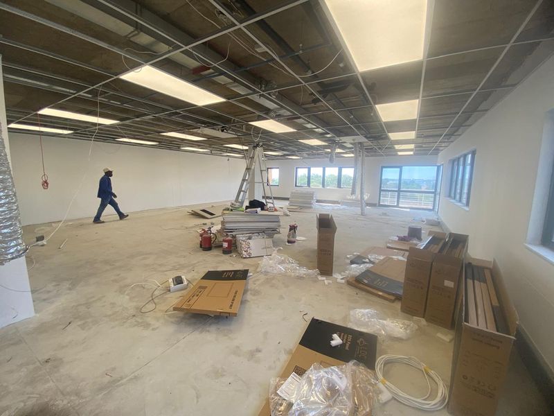 Spacious open-plan industrial office area currently under renovation, featuring a suspended ceiling grid, large windows, and concrete flooring.