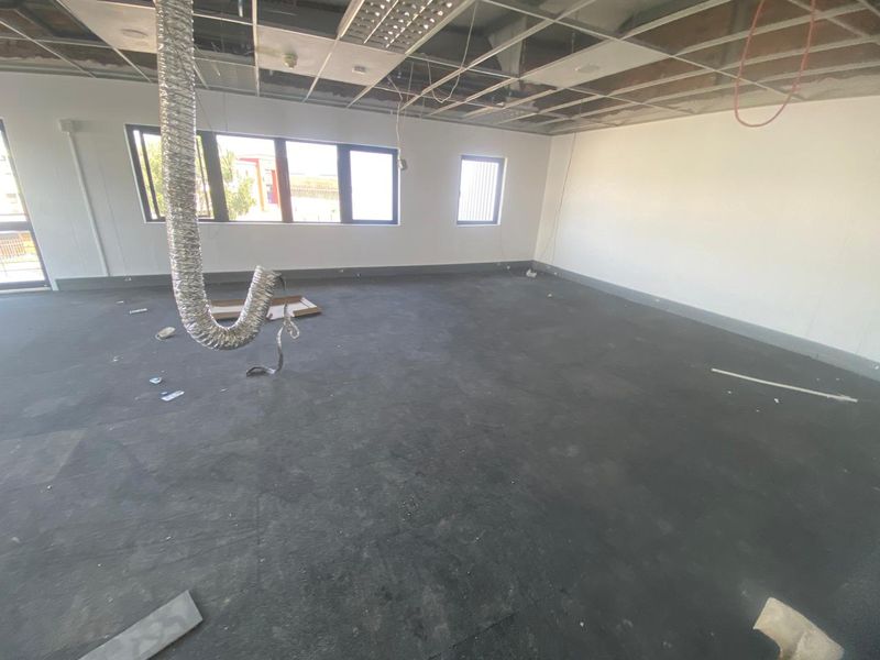 An open-plan industrial office space currently undergoing renovation, featuring exposed ceiling grid, ventilation ducting, and large windows providing natural light.