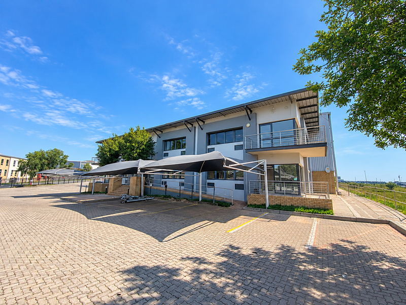 Modern industrial office building featuring a paved parking area with shade cloth carports, brick facade, and an upper-level balcony with steel railings.