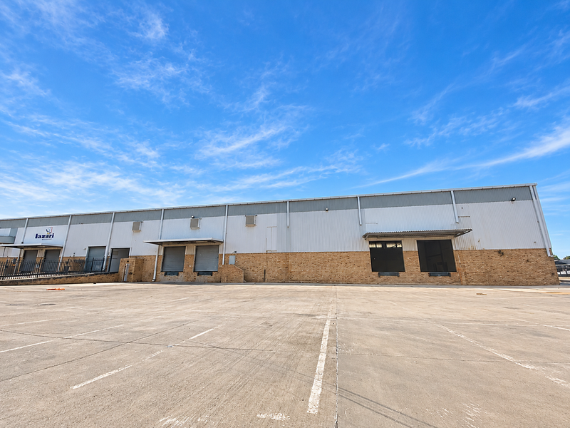 Industrial warehouse exterior featuring a large concrete yard, multiple loading docks with roller shutter doors, and a combination of brick and metal cladding.