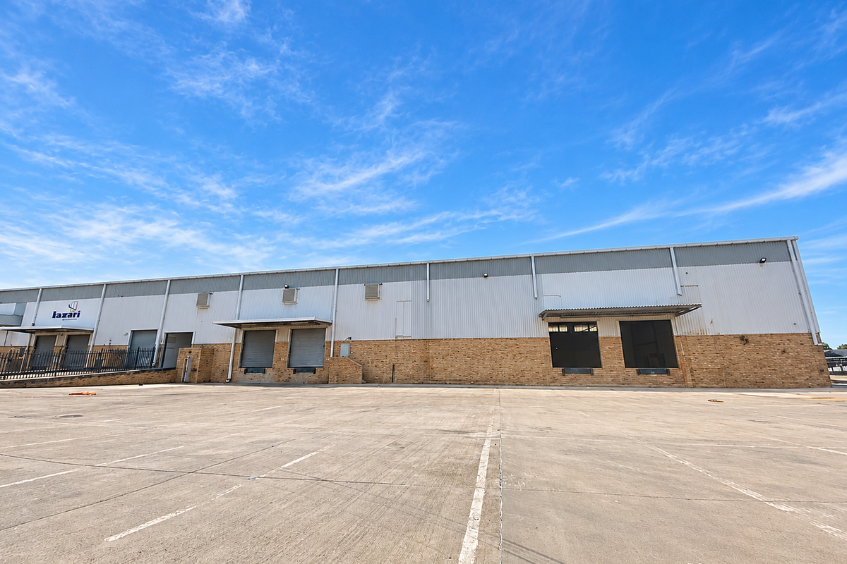 Industrial warehouse exterior featuring a large concrete yard, multiple loading docks with roller shutter doors, and a combination of brick and metal cladding.