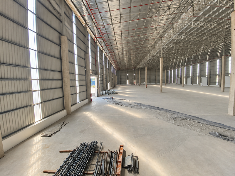 Expansive industrial warehouse space featuring high ceilings, concrete flooring, steel truss roofing, and natural light through translucent wall panels.