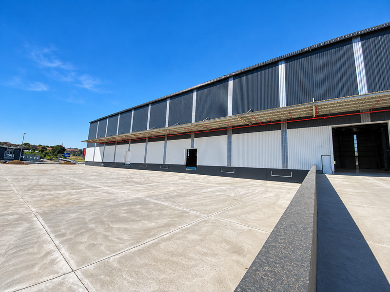 A modern industrial warehouse exterior featuring a large concrete loading yard, corrugated metal cladding, and multiple loading bay access points.