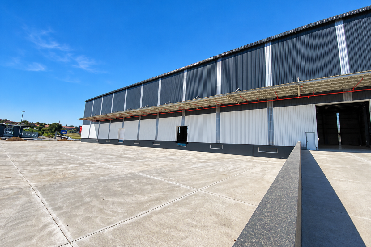 A modern industrial warehouse exterior featuring a large concrete loading yard, corrugated metal cladding, and multiple loading bay access points.