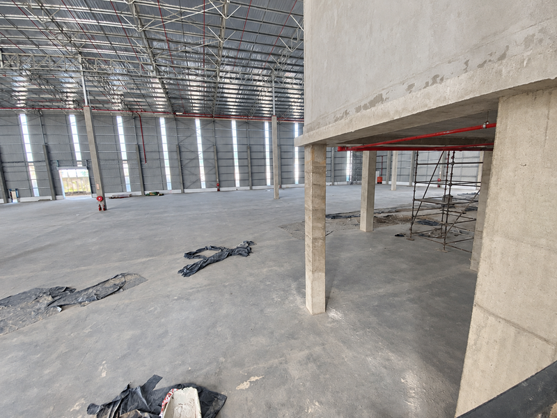 Spacious industrial warehouse interior featuring high ceilings with exposed steel trusses, concrete flooring, and fire sprinkler systems, currently in an unfinished construction state.