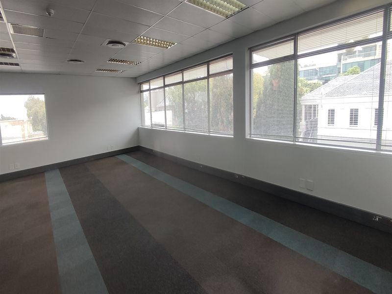 A spacious, empty office area featuring large windows with fitted blinds, suspended ceilings with recessed lighting, and carpeted flooring.