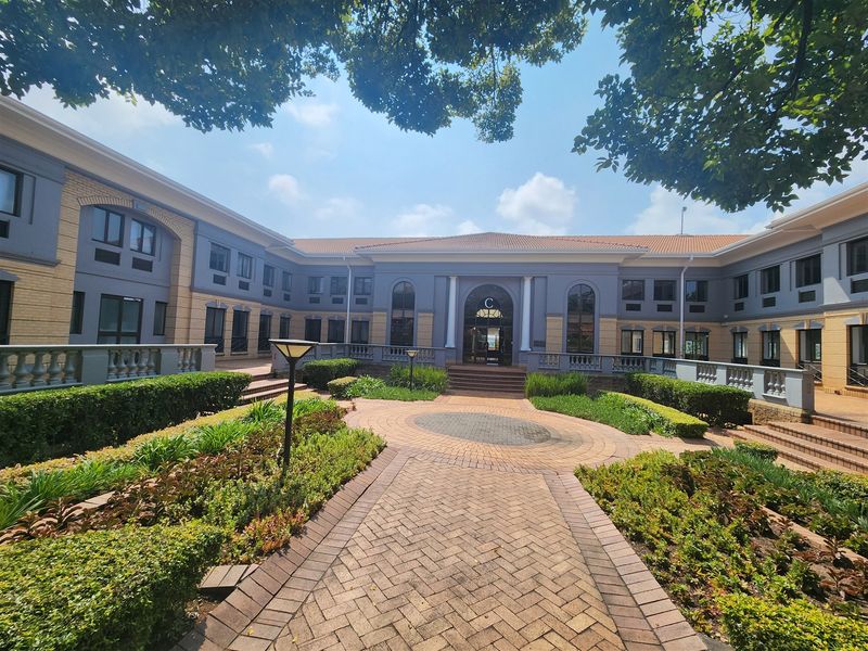 A well-maintained office building courtyard featuring paved walkways, landscaped garden beds, and classic architectural balustrades leading to the main entrance.