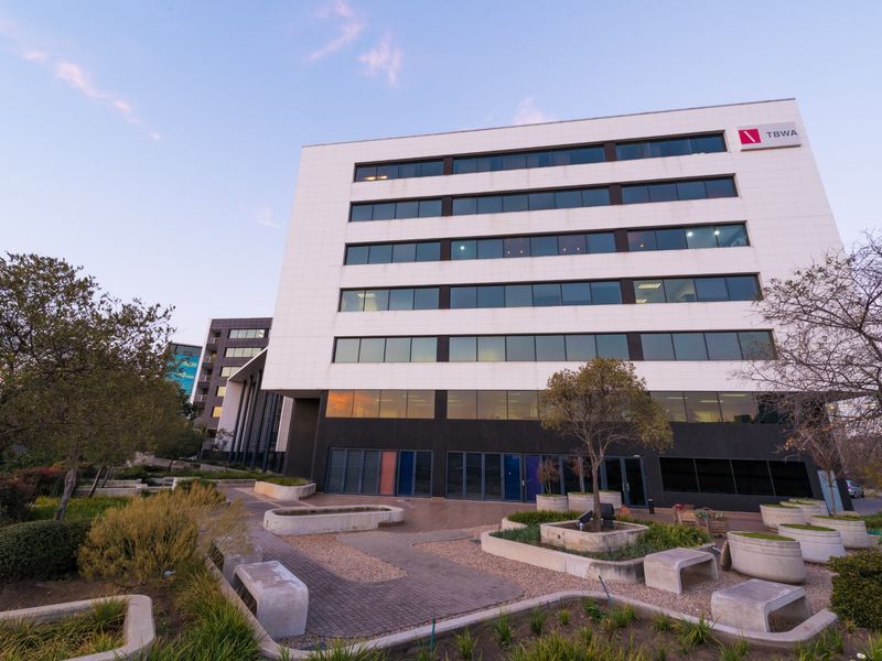 A modern multi-story office building featuring a clean white facade, extensive glass windows, and a landscaped paved courtyard with concrete seating areas.