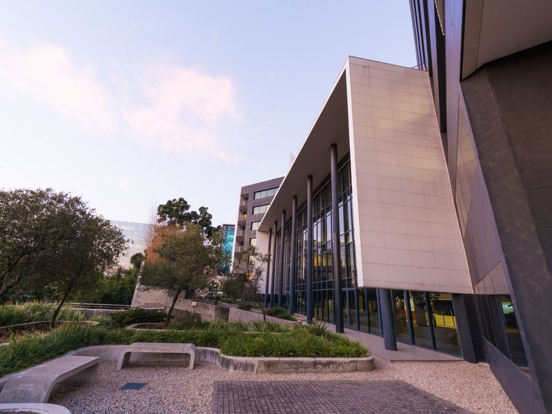 Modern office building exterior featuring a large glass curtain wall, structural columns, and a landscaped courtyard with concrete seating.