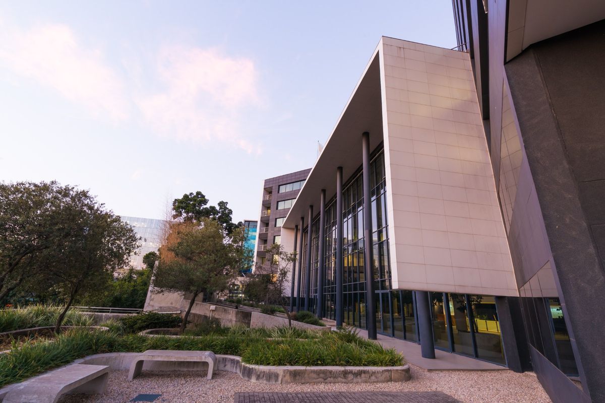 Modern office building exterior featuring a large glass curtain wall, structural columns, and a landscaped courtyard with concrete seating.