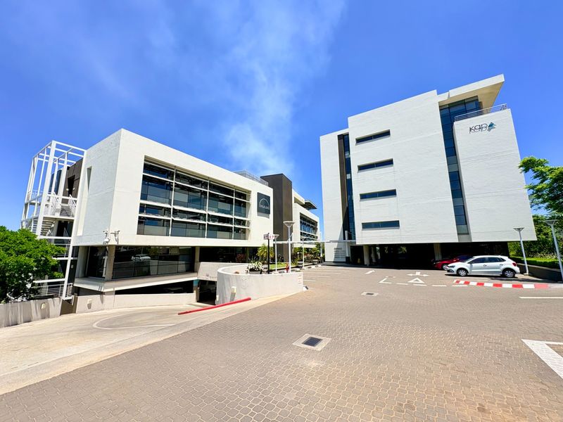 Modern multi-story office complex featuring contemporary white facades, glass curtain walls, and a paved driveway leading to an underground parking entrance.