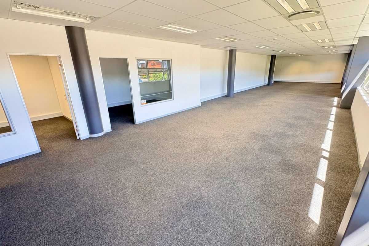 A spacious, open-plan office area featuring neutral carpeting, a suspended ceiling with recessed lighting, and structural columns, offering a clean and professional workspace.