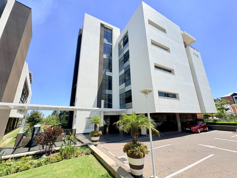 Modern multi-story office building exterior featuring a clean white facade, paved parking area, and well-maintained landscaped entrance.