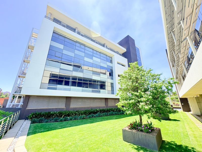 Modern multi-story office building featuring a glass curtain wall facade, external steel staircase, and a well-maintained landscaped garden area with concrete planters.