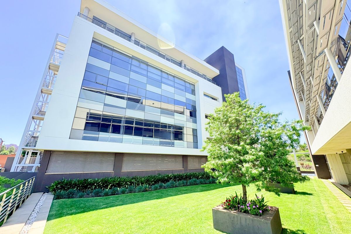 Modern multi-story office building featuring a glass curtain wall facade, external steel staircase, and a well-maintained landscaped garden area with concrete planters.