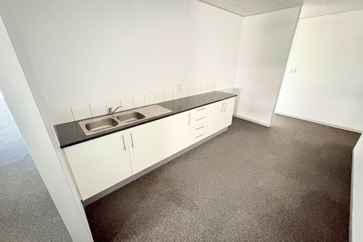 A clean, empty office kitchenette featuring white built-in cabinetry, a stainless steel double sink, dark countertop, and a tiled backsplash.