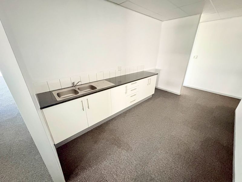 A clean, empty office kitchenette featuring white built-in cabinetry, a stainless steel double sink, dark countertop, and a tiled backsplash.